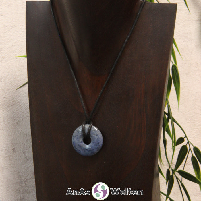 Das Bild zeigt einen Dumortierit Donut an einem Baumwollband. Der Edelstein hat eine dunkelblaue Grundfarbe mit hellblauen bis grau-blauen Flecken.