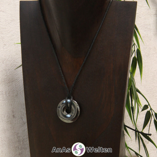 Das Bild zeigt einen Hämatit Donut an einem Baumwollband. Der Edelsteindonut ist silber-grau-metallen und hat eine glänzende und spiegelnde Oberfläche.