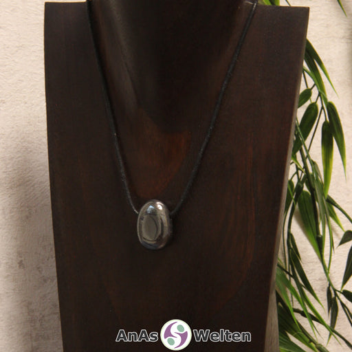 Ein gebohrter Hämatit Trommelstein an einem Baumwollband. Der Edelstein hat eine grau-metallene Oberfläche, die glänzt und spiegelt.