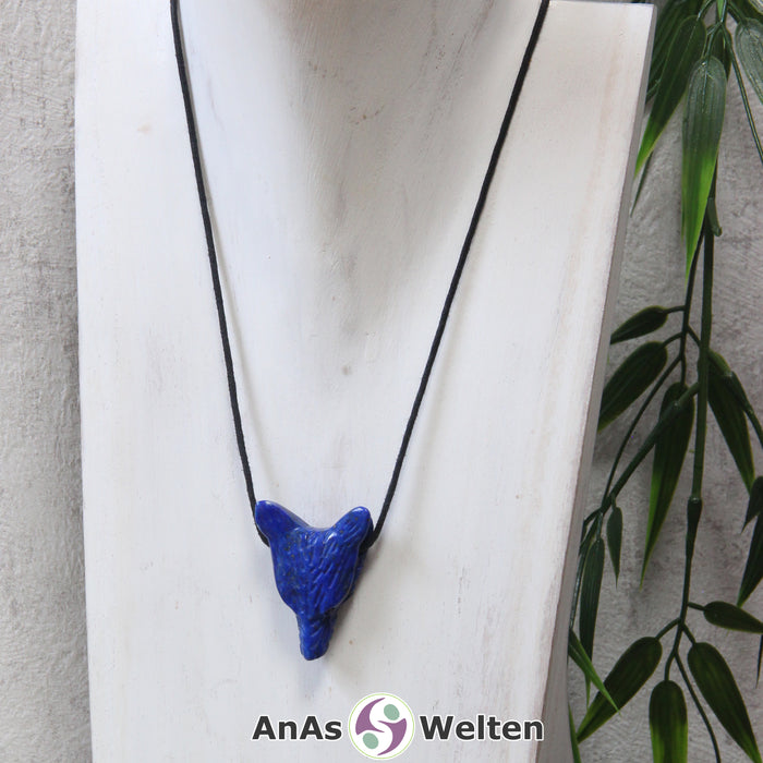 Das Bild zeigt einen Lapislazuli Wolf Anhänger an einem Baumwollband. Der Edelstein hat tiefblaue Farbe, die im Licht schwach golden glitzert. Das Fellmuster und die Nase sind durch kleine Kerben im Edelstein dargestellt. Die Augen sind kleine schwarze Perlen.