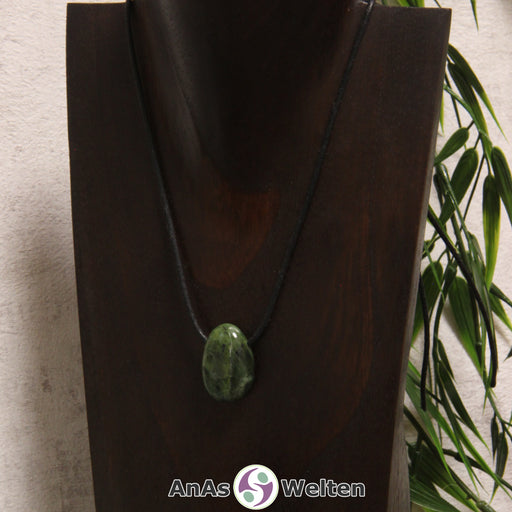 Ein gebohrter Nephrit Jade Trommelstein an einem Baumwollband. Der Edelstein hat eine Mischung aus dunkel- und Hellgrün. An einigen Stellen ist er von schwarzen Punkten durchzogen.