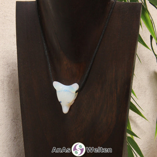 Das Bild zeigt einen Opalglas Wolf Anhänger an einem Baumwollband. Der Edelstein hat eine schwach durchsichtige weiße Grundfarbe und schimmert blau und rot im Licht. Das Fellmuster und die Nase sind durch kleine Kerben im Edelstein dargestellt. Die Augen sind kleine schwarze Perlen.