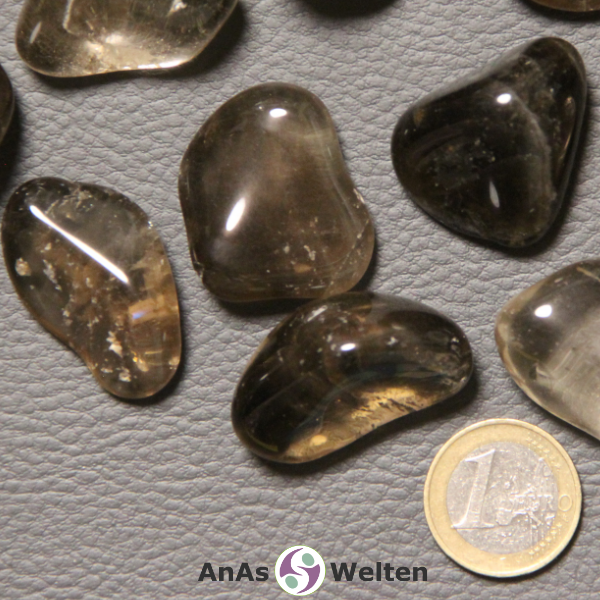 Das Produktbild zeigt einen Rauchquarz Trommelstein anhand mehrerer Beispielsteine. Die Edelsteine sind grau-braun und größtenteils durchsichtig, wodurch man die Einschlüsse und Risse in ihrem Inneren gut sehen kann.
