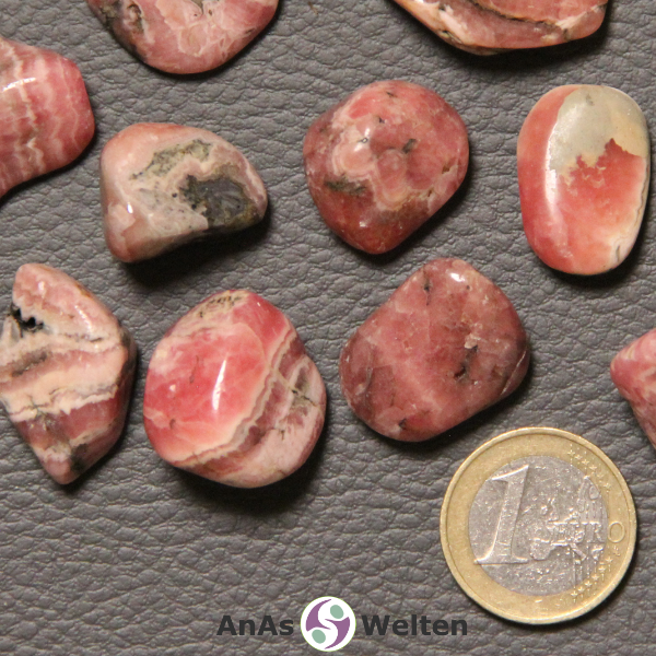 Das Bild zeigt einen Rhodochrosit Trommelstein anhand mehrerer Beispielsteine. Die Edelsteine haben allesamt eine Grundfarbe aus Lachs und Beige. Außerdem haben die meisten von ihnen grau-schwarze Einschlüsse.