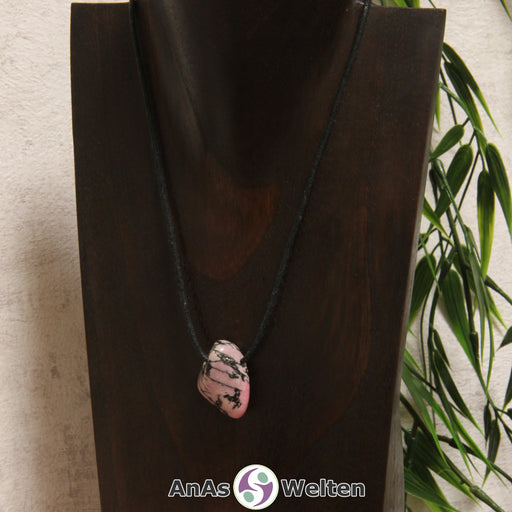 Ein gebohrter Rhodonit Trommelstein an einem Baumwollband. Der Edelstein hat eine rot-beige Farbe und schwarze Einschlüsse.