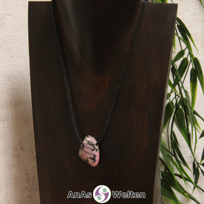 Ein gebohrter Rhodonit Trommelstein an einem Baumwollband. Der Edelstein hat eine rot-beige Farbe und schwarze Einschlüsse.