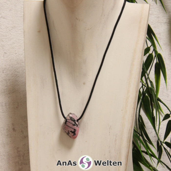 Ein rosa-schwarze Rhodonit gebohrter Trommelstein an einem schwarzen Baumwollband, der um eine helle Büste gehängt wurde.