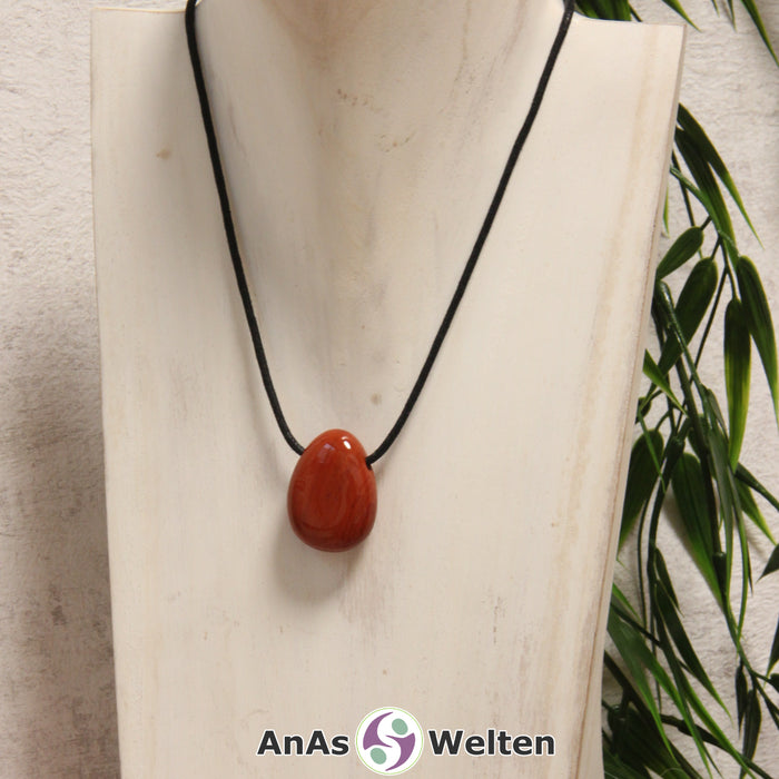 Ein Roter Jaspis gebohrter Trommelstein an einem schwarzen Baumwollband, der um eine helle Büste gehängt wurde.