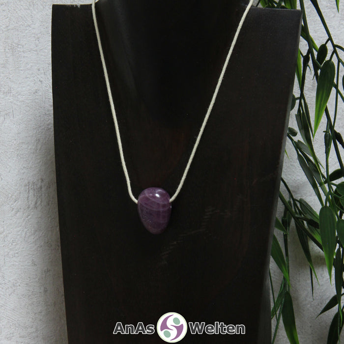 Ein lila-roter Rubin gebohrter Trommelstein an einem beigen Baumwollband, der um eine dunkle Büste gehängt wurde.