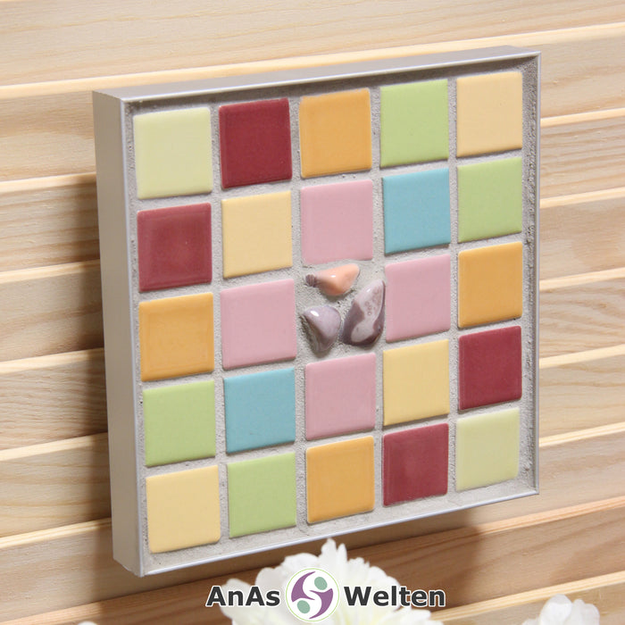 Das Bild zeigt ein Sator Quadrat Mosaik in Pastellfarben mit einem Aluminiumrahmen. Dort, wo im Sator Quadrat die Buchstaben stehen, sind hier viereckige Fliesen zu sehen, die je eine Farbe pro unterschiedlichen Buchstaben haben. In der Mitte, wo normalerweise das N steht, sind drei rosa Achate in den Fugenmörtel eingesetzt.