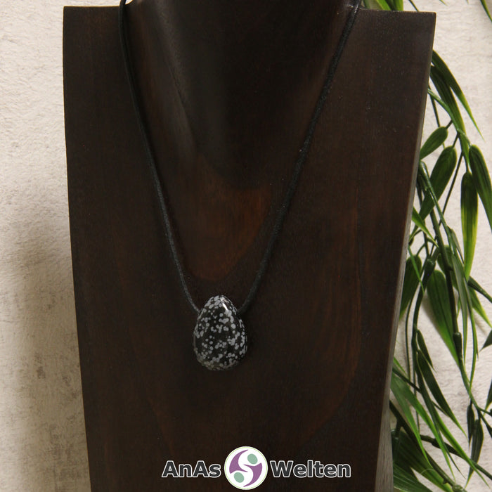 Ein schwarz-weiße Schneeflocken-Obsidian gebohrter Trommelstein an einem schwarzen Baumwollband, der um eine dunkle Büste gehängt wurde.