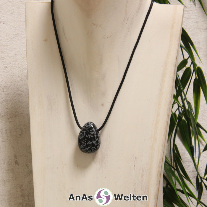 Ein gebohrter Schneeflocken-Obsidian Trommelstein an einem Baumwollband. Der Edelstein hat eine schwarze Grundfarbe mit vielen kleinen grau-weißen Flecken.
