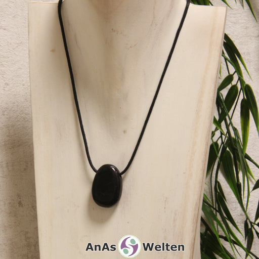 Ein gebohrter Schungit Trommelstein an einem Baumwollband. Der Edelstein hat eine schwarze Farbe mit einem leicht matten Glanz.