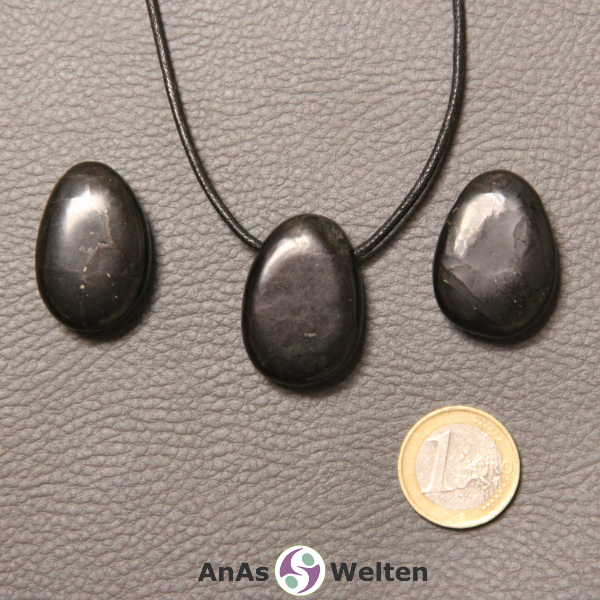 Drei schwarze Schungit gebohrte Trommelsteine nebeneinander. Einer befindet sich an einem schwarzen gewachsten Baumwollband.