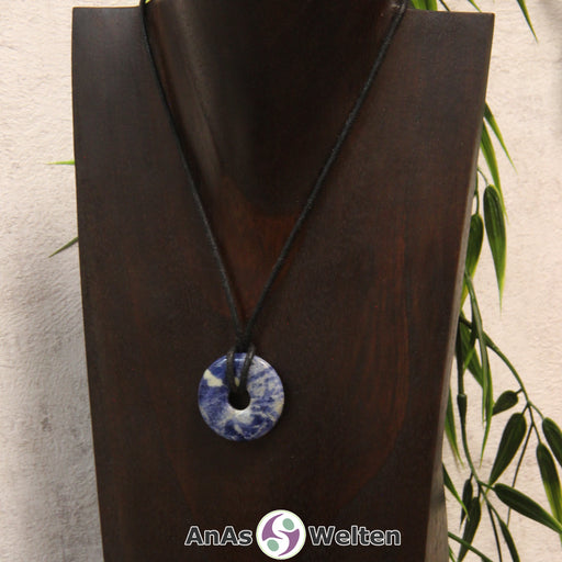 Das Bild zeigt einen Sodalith Donut an einem Baumwollband. Der Edelstein hat eine blau-weiße Musterung. Die beiden Farben sind dabei recht gleichmäßig verteilt.