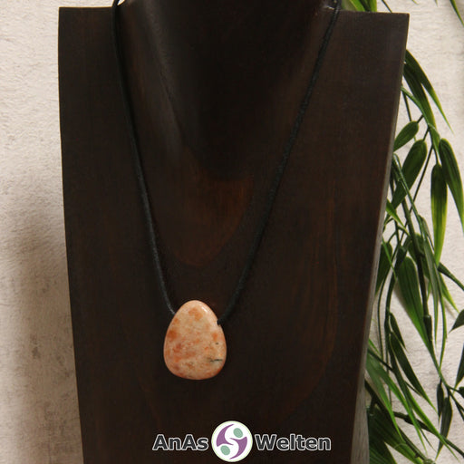 Ein gebohrter Sonnenstein Trommelstein an einem Baumwollband. Der Edelstein hat eine orange-beige Farbe, deren Muster ein wenig an Flammen erinnert.