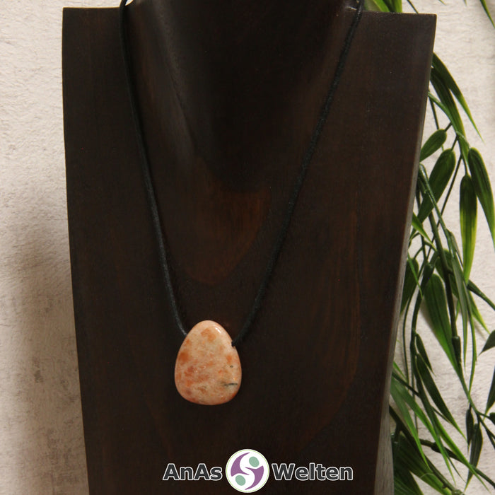 Ein gebohrter Sonnenstein Trommelstein an einem Baumwollband. Der Edelstein hat eine orange-beige Farbe, deren Muster ein wenig an Flammen erinnert.