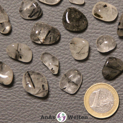 Das Bild zeigt einen Turmalinquarz Trommelstein anhand mehrerer Beispielsteine. Die Edelsteine haben allesamt eine durchsichtige, milchig-weiße oder halbdurchsichtig graue Farbe. Außerdem verfügen sie über meist strichartige schwarze Einschlüsse, die bei den meisten Steinen gut sichtbar sind.