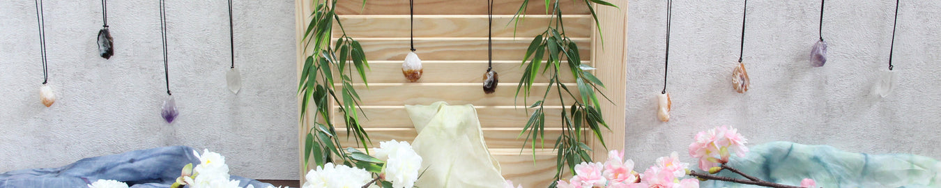 Das Bild zeigt diverse Edelstein Rohstein Ketten an einer weißen Wand. Als Deko liegen bunte Tücher und Kirschblüten auf dem Boden.