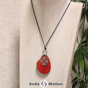 Das Produktbild für die 1. Chakra (Wurzelchakra) Symbol Kette rot zeigt eine rot gefärbte Tagua-Nuss-Scheibe an einem schwarzen Baumwollband, vor der ein silberfarbener Anhänger mit dem Chakrasymbol befestigt ist. Die Nuss-Scheibe hat einen dunklen Rand und eine glänzende Oberfläche. Direkt darüber befindet sich eine Bergkristallkugel.