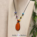 Das Produktbild für die 2. Chakra (Sakralchakra) Kette bunt zeigt eine orange gefärbte Tagua-Nuss-Scheibe an einem schwarzen Baumwollband. Darüber befinden sich mit Knoten befestigte Taugua-Nuss-Splitter in den Farben Rot, Gelb, Lila, Grün, Blau und Hellblau. Die Nuss-Scheibe hat einen dunklen Rand und eine glänzende Oberfläche.