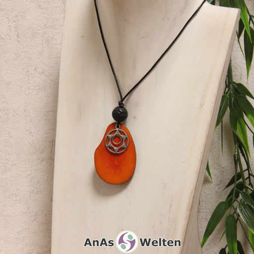 Das Produktbild für die 2. Chakra (Sakralchakra) Symbol Kette orange zeigt eine orange gefärbte Tagua-Nuss-Scheibe an einem schwarzen Baumwollband, vor der ein silberfarbener Anhänger mit dem Chakrasymbol befestigt ist. Die Nuss-Scheibe hat einen dunklen Rand und eine glänzende Oberfläche. Direkt darüber befindet sich eine Lavakugel.