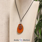 Das Produktbild für die 2. Chakra (Sakralchakra) Symbol Kette orange zeigt eine orange gefärbte Tagua-Nuss-Scheibe an einem schwarzen Baumwollband, vor der ein silberfarbener Anhänger mit dem Chakrasymbol befestigt ist. Die Nuss-Scheibe hat einen dunklen Rand und eine glänzende Oberfläche. Direkt darüber befindet sich eine Bergkristallkugel.