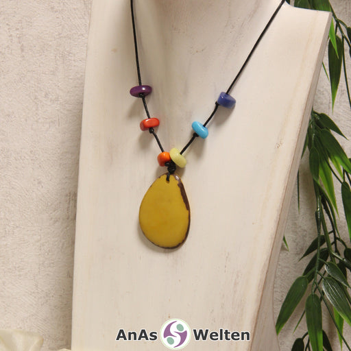 Das Produktbild für die 3. Chakra (Solarplexuschakra) Kette bunt zeigt eine gelb gefärbte Tagua-Nuss-Scheibe an einem schwarzen Baumwollband. Darüber befinden sich mit Knoten befestigte Taugua-Nuss-Splitter in den Farben Orange, Gelb, Rot, Hellblau, Lila und Blau. Die Nuss-Scheibe hat einen dunklen Rand und eine glänzende Oberfläche.