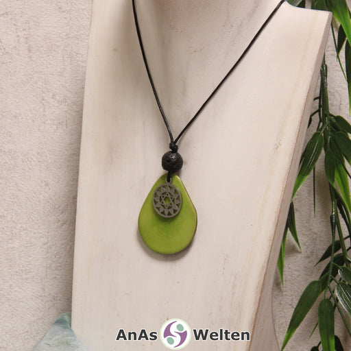 Das Produktbild für die 4. Chakra (Herzchakra) Symbol Kette grün zeigt eine apfelgrün gefärbte Tagua-Nuss-Scheibe an einem schwarzen Baumwollband, vor der ein silberfarbener Anhänger mit dem Chakrasymbol befestigt ist. Die Nuss-Scheibe hat einen dunklen Rand und eine glänzende Oberfläche. Direkt darüber befindet sich eine Lavakugel.