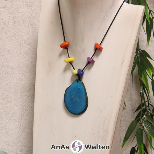 Das Produktbild für die 5. Chakra (Halschakra) Kette bunt zeigt eine hellblau gefärbte Tagua-Nuss-Scheibe an einem schwarzen Baumwollband. Darüber befinden sich mit Knoten befestigte Taugua-Nuss-Splitter in den Farben Grün, Blau, Gelb, Lila, Orange und Rot. Die Nuss-Scheibe hat einen dunklen Rand und eine glänzende Oberfläche.