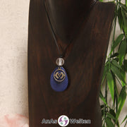 Eine blaue Tagua-Nuss Chakra Kette mit silberfarbenem 6. Chakra Symbol und Bergkristallkugel an einer dunklen Büste.