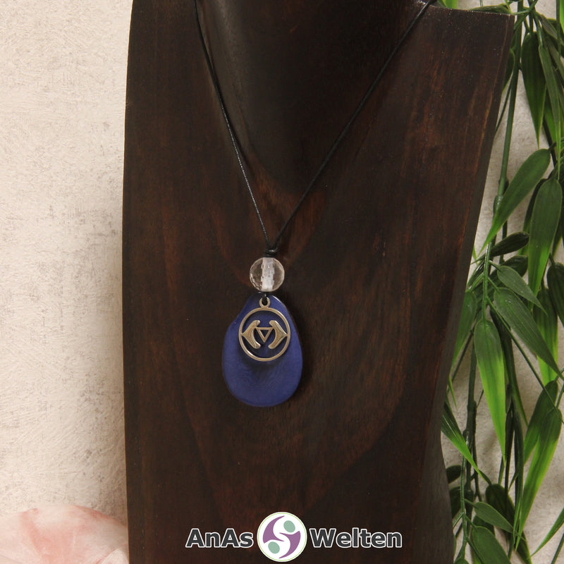Eine blaue Tagua-Nuss Chakra Kette mit silberfarbenem 6. Chakra Symbol und Bergkristallkugel an einer dunklen Büste.