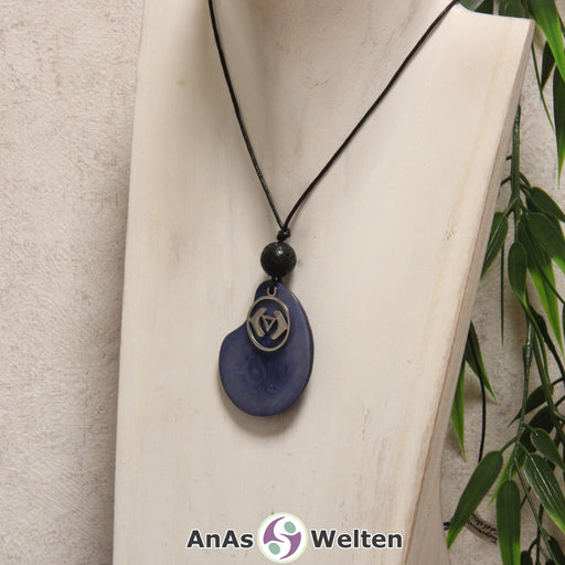 Das Produktbild für die 6. Chakra (Stirnchakra) Symbol Kette blau zeigt eine blau gefärbte Tagua-Nuss-Scheibe an einem schwarzen Baumwollband, vor der ein silberfarbener Anhänger mit dem Chakrasymbol befestigt ist. Die Nuss-Scheibe hat einen dunklen Rand und eine glänzende Oberfläche. Direkt darüber befindet sich eine Lavakugel.