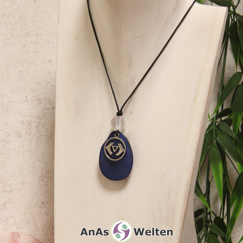 Das Produktbild für die 6. Chakra (Stirnchakra) Symbol Kette blau zeigt eine blau gefärbte Tagua-Nuss-Scheibe an einem schwarzen Baumwollband, vor der ein silberfarbener Anhänger mit dem Chakrasymbol befestigt ist. Die Nuss-Scheibe hat einen dunklen Rand und eine glänzende Oberfläche. Direkt darüber befindet sich eine Bergkristallkugel.