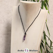 Amethyst Stift-Anhänger klein mit Kette: Das Bild zeigt einen Amethyst Stift-Anhänger klein mit Kette. Der Stift-Anhänger ist lila mit einem weißen Einschluss. Am oberen Ende verbindet ein silberfarbenes Element den Edelstein mit der Stoffkordel.