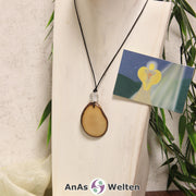 Das Produktbild für die Engel Kette Erzengel Azrael mit Karte zeigt eine in einem blassen Gelb gefärbte Tagua-Nuss-Scheibe an einem schwarzen Baumwollband. Die Nuss-Scheibe hat einen dunklen Rand und eine glänzende Oberfläche. Direkt darüber befindet sich eine facettierte Bergkristallkugel. Rechts neben der Kette sieht man außerdem die Erzengel Azrael Karte, die den Engel als abstraktes Bild zeigt.