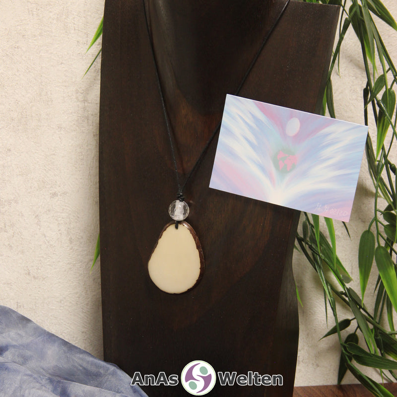 Das Produktbild für die Engel Kette Erzengel Gabriel mit Karte zeigt eine Tagua-Nuss-Scheibe in ihrem natürlichen Cremeweiß an einem schwarzen Baumwollband. Die Nuss-Scheibe hat einen dunklen Rand und eine glänzende Oberfläche. Direkt darüber befindet sich eine facettierte Bergkristallkugel. Rechts neben der Kette sieht man außerdem die Erzengel Gabriel Karte, die den Engel als abstraktes Bild zeigt.