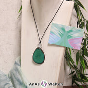 Das Produktbild für die Engel Kette Erzengel Haniel mit Karte zeigt eine in einem kräftigen Grün gefärbte Tagua-Nuss-Scheibe an einem schwarzen Baumwollband. Die Nuss-Scheibe hat einen dunklen Rand und eine glänzende Oberfläche. Direkt darüber befindet sich eine facettierte Bergkristallkugel. Rechts neben der Kette sieht man außerdem die Erzengel Haniel Karte, die den Engel als abstraktes Bild zeigt.