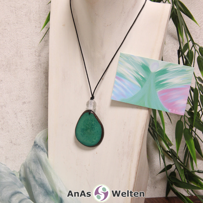 Das Produktbild für die Engel Kette Erzengel Haniel mit Karte zeigt eine in einem kräftigen Grün gefärbte Tagua-Nuss-Scheibe an einem schwarzen Baumwollband. Die Nuss-Scheibe hat einen dunklen Rand und eine glänzende Oberfläche. Direkt darüber befindet sich eine facettierte Bergkristallkugel. Rechts neben der Kette sieht man außerdem die Erzengel Haniel Karte, die den Engel als abstraktes Bild zeigt.