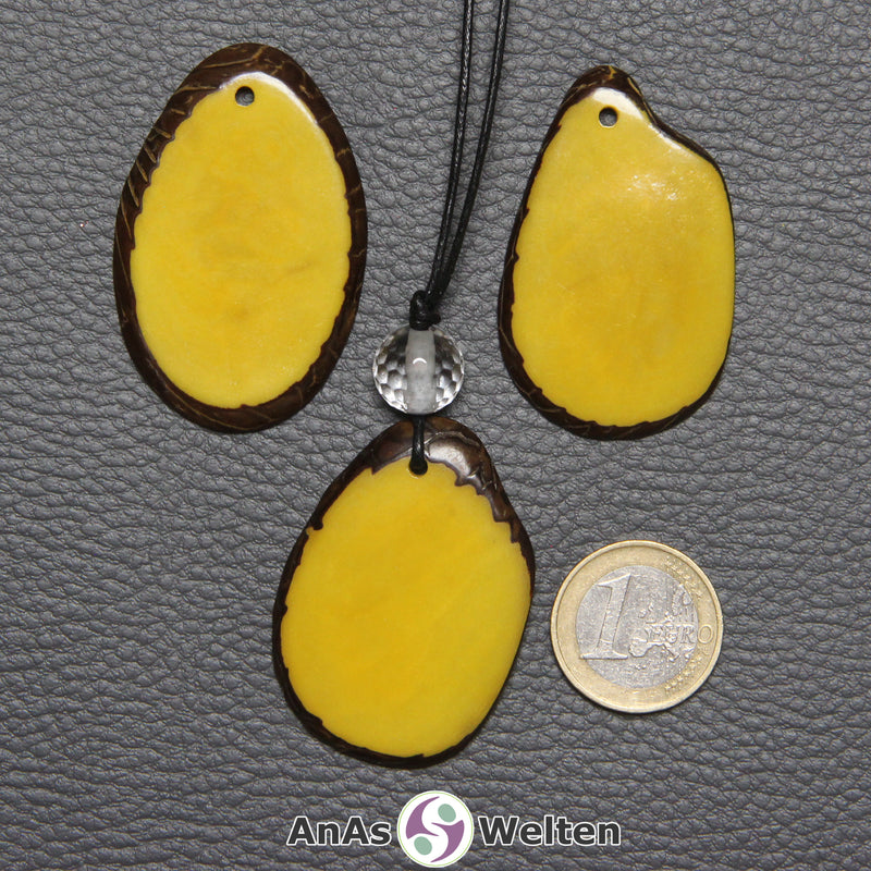 Drei Tagua-Nuss-Scheiben in Gelb. Die mittlere ist mit einer Bergkristallkugel an ein schwarzes Baumwollband gebunden.