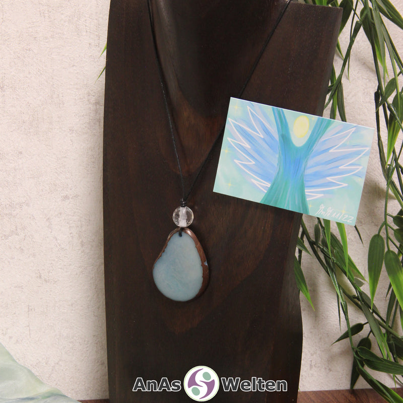 Das Produktbild für die Engel Kette Erzengel Raguel mit Karte zeigt eine in einem blassen Blau gefärbte Tagua-Nuss-Scheibe an einem schwarzen Baumwollband. Die Nuss-Scheibe hat einen dunklen Rand und eine glänzende Oberfläche. Direkt darüber befindet sich eine facettierte Bergkristallkugel. Rechts neben der Kette sieht man außerdem die Erzengel Raguel Karte, die den Engel als abstraktes Bild zeigt.