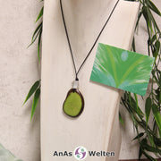 Das Produktbild für die Engel Kette Erzengel Raphael mit Karte zeigt eine in einem kräftigen Grün gefärbte Tagua-Nuss-Scheibe an einem schwarzen Baumwollband. Die Nuss-Scheibe hat einen dunklen Rand und eine glänzende Oberfläche. Direkt darüber befindet sich eine facettierte Bergkristallkugel. Rechts neben der Kette sieht man außerdem die Erzengel Raphael Karte, die den Engel als abstraktes Bild zeigt.