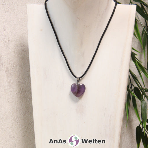 Das Bild zeigt einen Herzanhänger Amethyst-Herz an einem schwarzen Baumwollband. Der Edelstein wurde in eine Herzform geschliffen und verfügt über eine silberfarbene Öse an der Oberseite. Er hat verschiedene Lilatöne und ist leicht durchsichtig, sodass man feine Risse in seinem Inneren erkennen kann, die ihm seine Struktur verleihen.