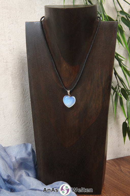 Das Bild zeigt einen Herzanhänger Opalglas-Herz an einem schwarzen Baumwollband. Der Edelstein wurde in eine Herzform geschliffen und verfügt über eine silberfarbene Öse an der Oberseite. Er hat eine halbdurchsichtige weiß-bläuliche Farbe und schimmert rot und blau.