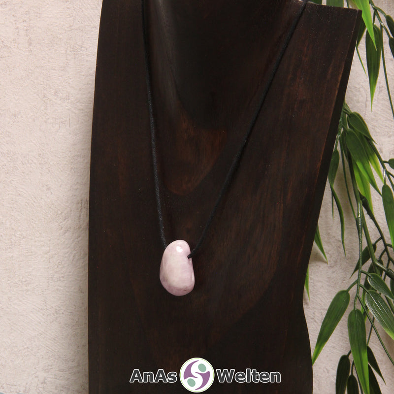 Ein gebohrter Kunzit Trommelstein an einem Baumwollband. Der Edelstein hat eine geschliffene, wenn auch unebene Form und eine zarte Rosafarbe.