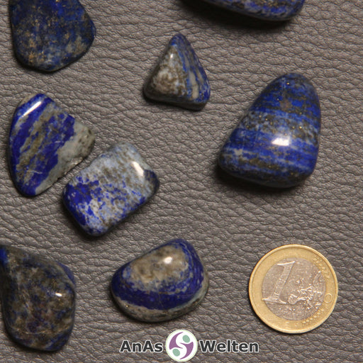 Das Produktbild zeigt einen Lapislazuli Trommelstein anhand von mehreren Beispielsteinen. Die Edelsteine haben einen tiefblauen Grundton und sind von gelegentlichem Grau, Braun und/oder Weiß durchzogen.