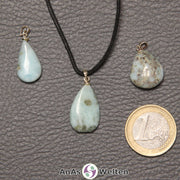 Drei blaue Larimar Steine mit Öse nebeneinander. Einer befindet sich an einem schwarzen gewachsten Baumwollband.