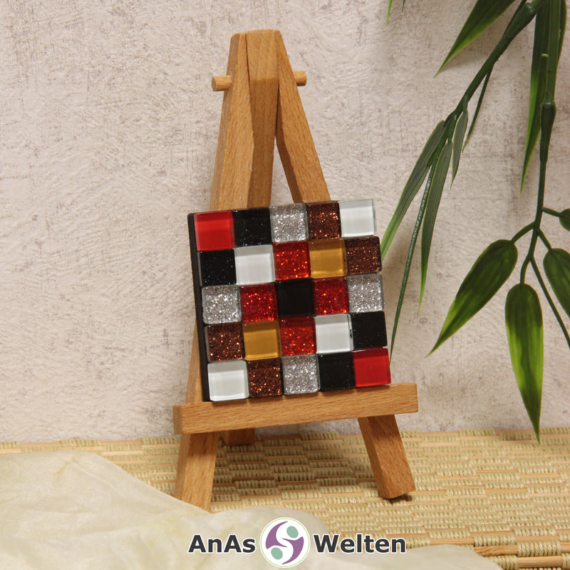 Das Bild zeigt ein Sator Quadrat Mosaik ohne Rahmen. Die Fliesen sind Glasfliesen in zwei verschiedenen Rottönen, zwei verschieden Schwarz, Silber, Bronze, Weiß und Braun. Einige von ihnen glitzern. Jeder Buchstabe auf dem Sator Quadrat wird durch eine andere Fliesenfarbe dargestellt.