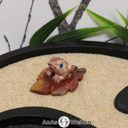 Das Bild zeigt ein Speckstein Frosch auf Blatt. Die Specksteinfigur stellt einen Frosch da, der auf einem Laubblatt sitzt. Der Frosch ist auf das Blatt geklebt, da sie aus unterschiedlichem Speckstein bestehen, damit sie unterschiedlich aussehen. Außerdem sind die Augen aus einem anderen Material, sodass sie besser zur Geltung kommen. Der Mund, die Zehen und das Blatt sind mit Kerben in den Stein gearbeitet.