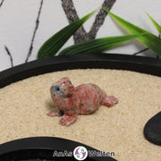 Das Bild zeigt eine Speckstein Robbe klein. Die Specksteinfigur stellt eine Robbe oder einen Seehund dar. Sie liegt auf dem Bauch, ist auf die vorderen Flossen gestützt und hat den Kopf zur Seite gerichtet. Die Augen sind aus einem anderen Material, damit sie besser zur Geltung kommen. Die Details an den Flossen und der Schnauze sind durch Einkerbungen im Stein dargestellt.